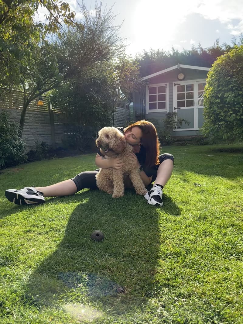 The groomer sitting on a sunlit lawn hugging a cream cockapoo